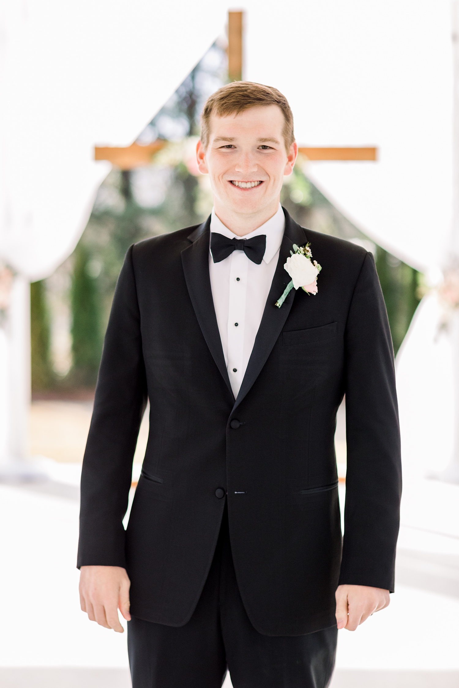 groom in classic tux poses in front of wooden cross