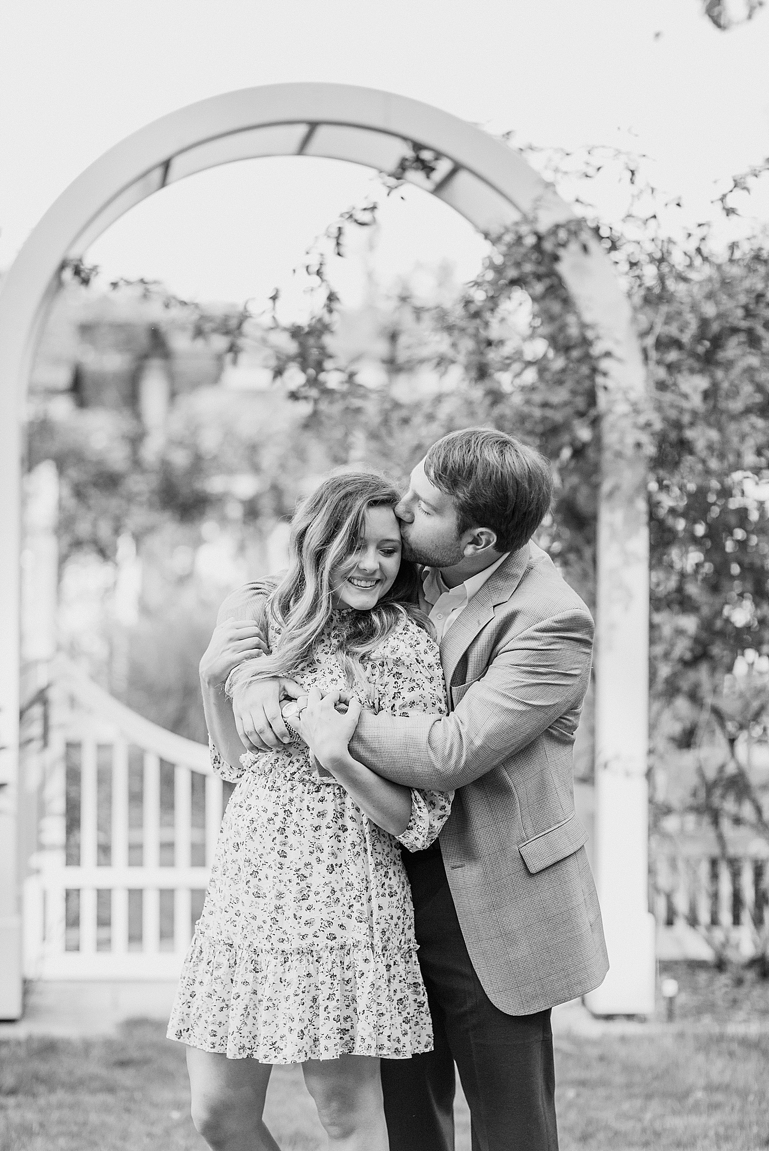 groom hugs bride from behind near white arch in Birmingham Botanical Gardens