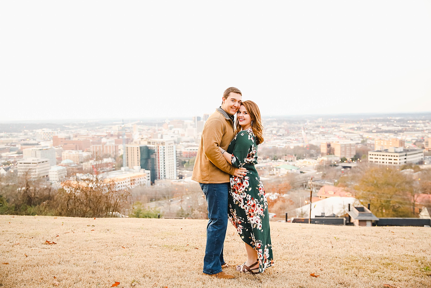 couple lean in on hill top with city down below during Downtown Birmingham AL Engagement Portraits