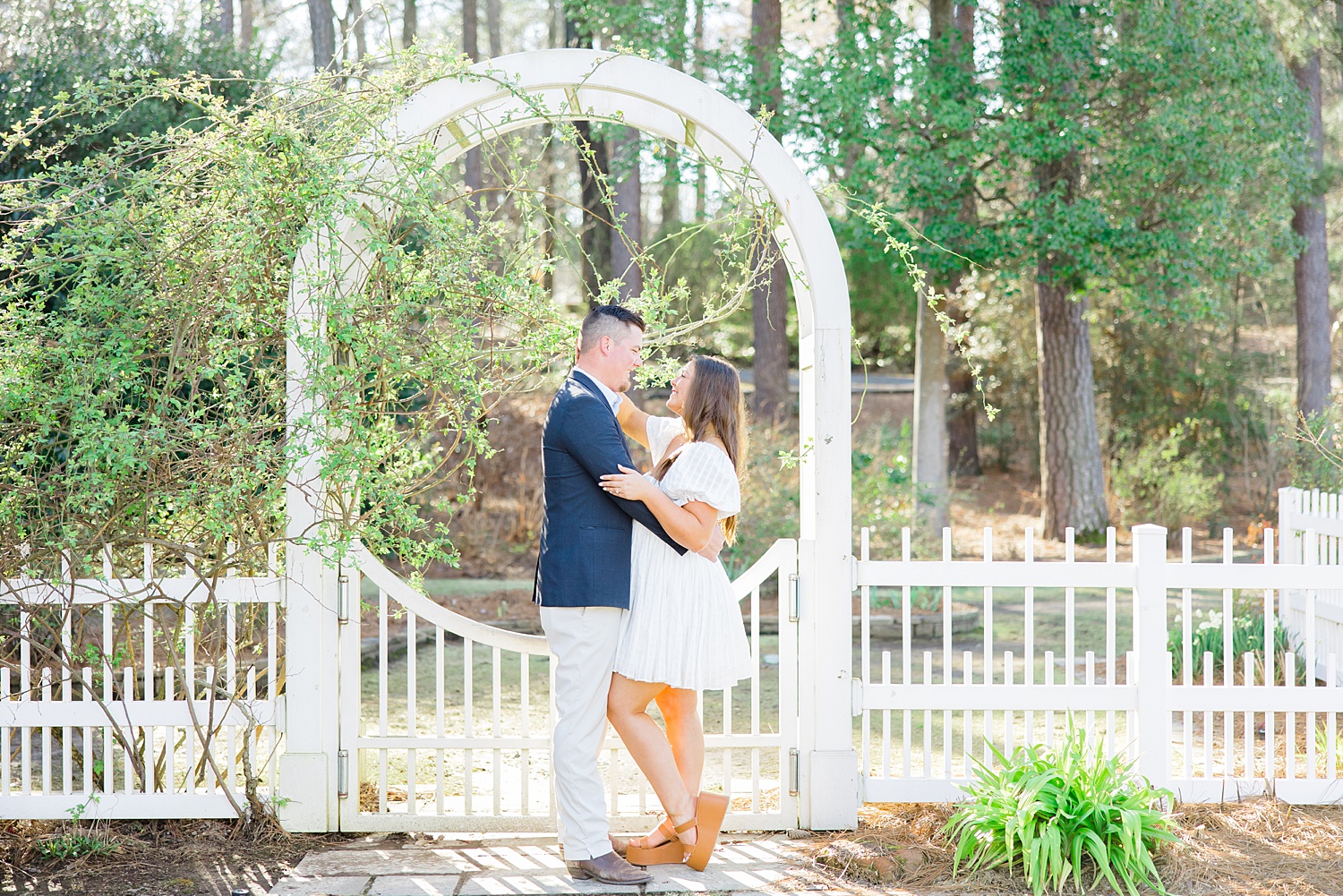 engaged couple stand under white arch in garden
