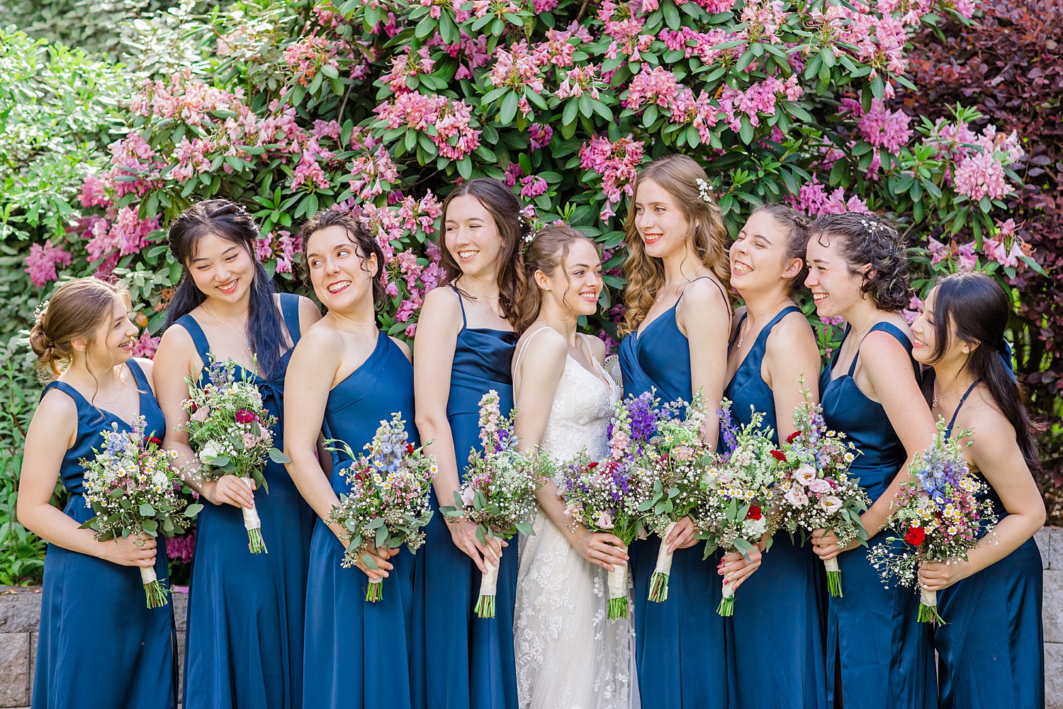 bride with bridesmaids in dark blue satin dresses holding garden inspired wedding flowers