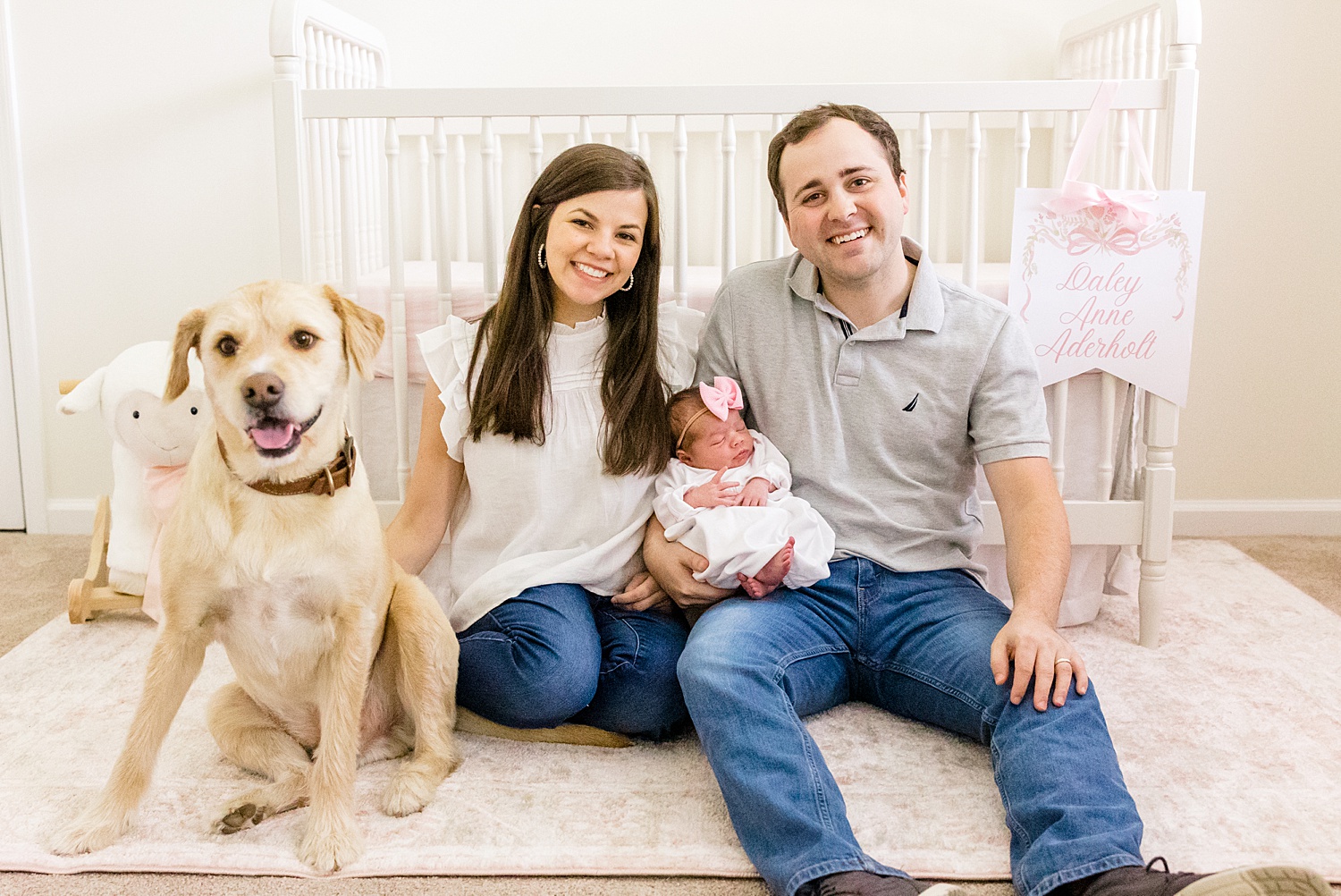 parents sit on nursery floor holding newborn girl with their family dog nearby