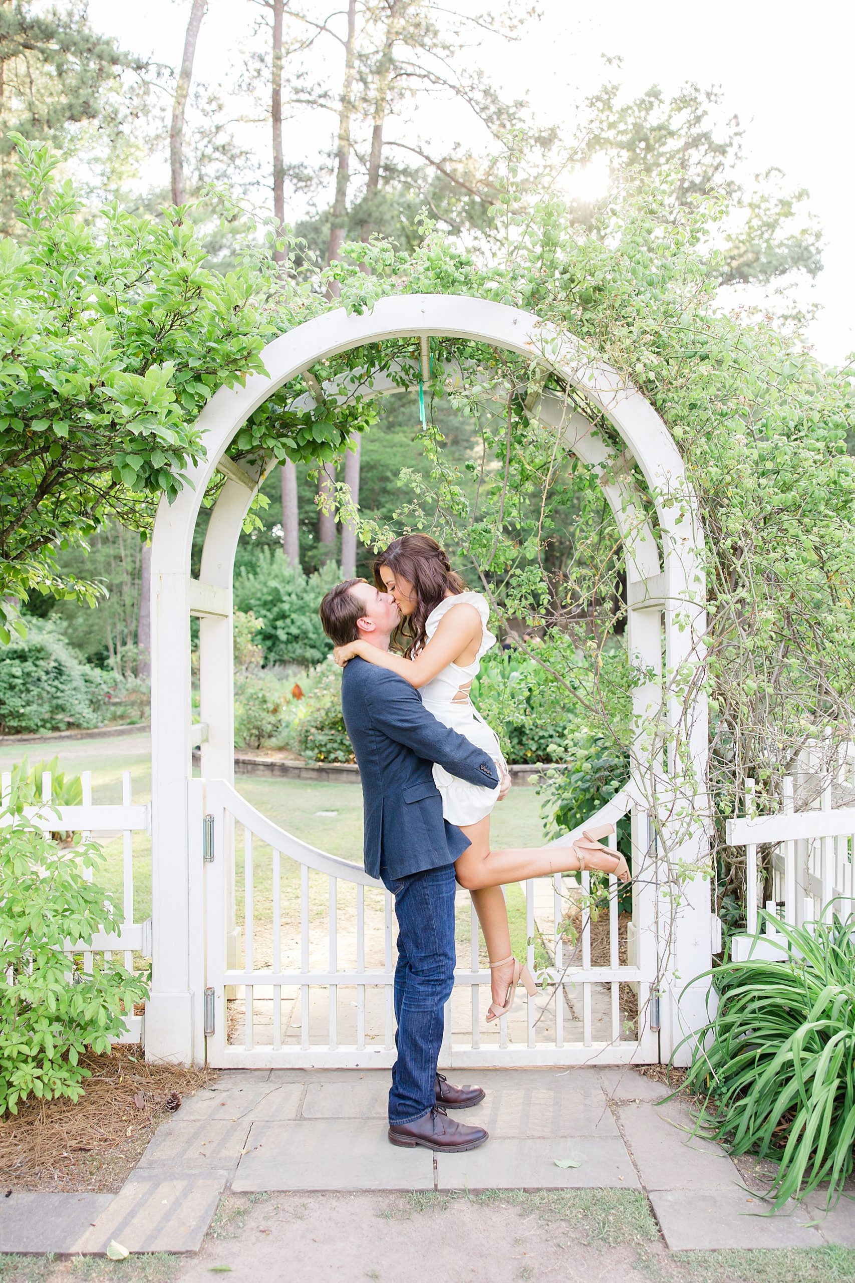 man lifts his fiance up under white arch in the garden